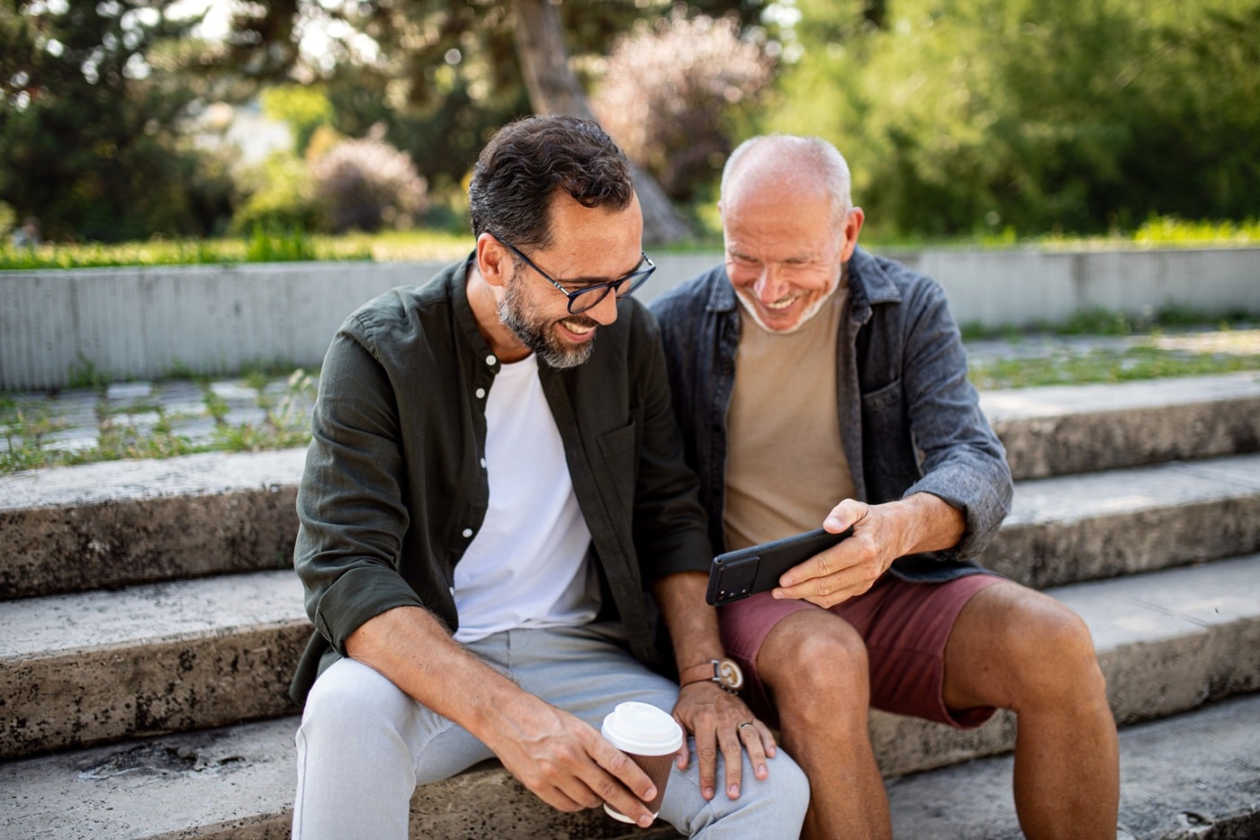 Two men sitting on outdoor steps, one holding a smartphone while they both look at the screen, engaged in conversation.